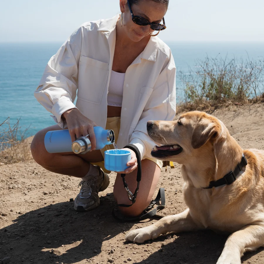 Woman pouring water from a light blue bottle into a light blue bowl for a dog on a dirt path by the ocean